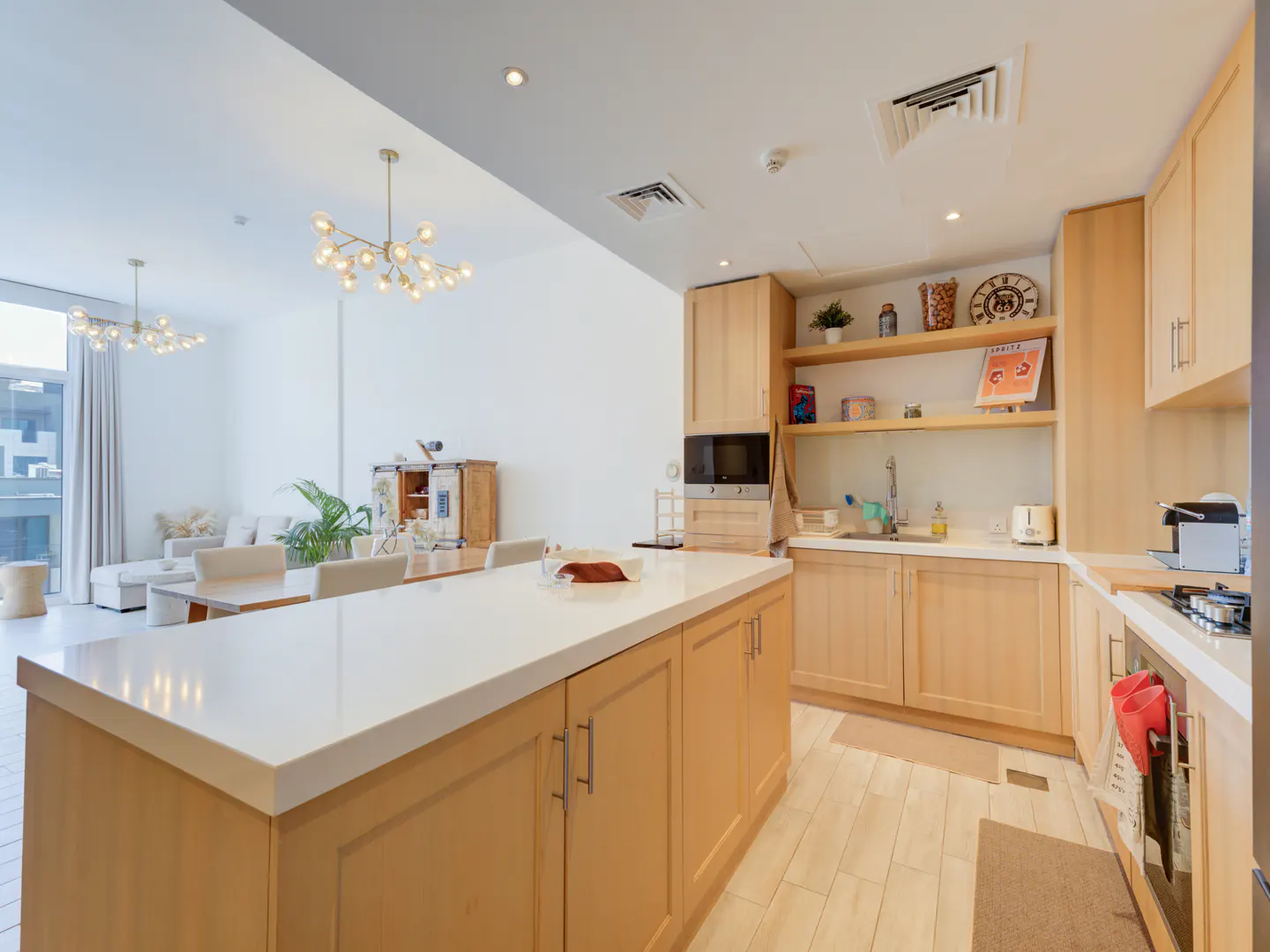 Bright, open-concept kitchen and living area. Light wood cabinets, white countertops, island, and modern light fixtures. Sofa and dining table visible.