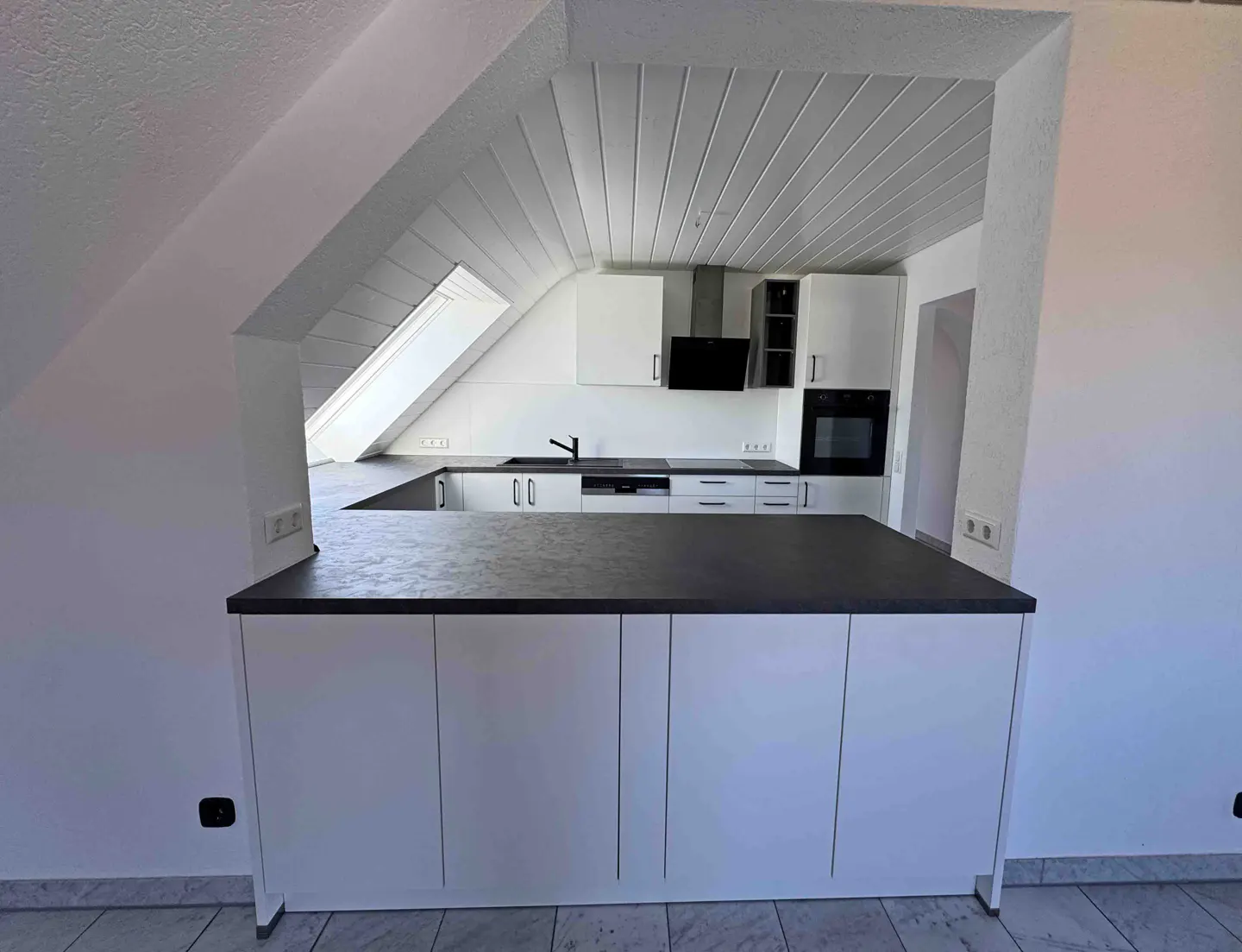 Modern attic kitchen with white cabinets, dark countertops, a skylight, and a built-in oven under a sloped white ceiling.