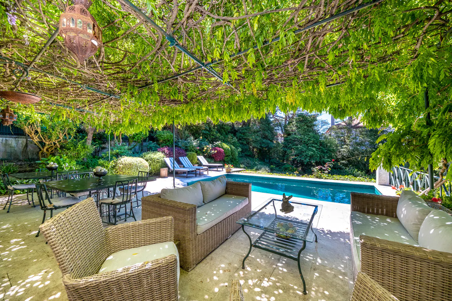 Outdoor patio with wicker furniture, a pool, and a vine-covered pergola. Lounge chairs and lush greenery in the background.
