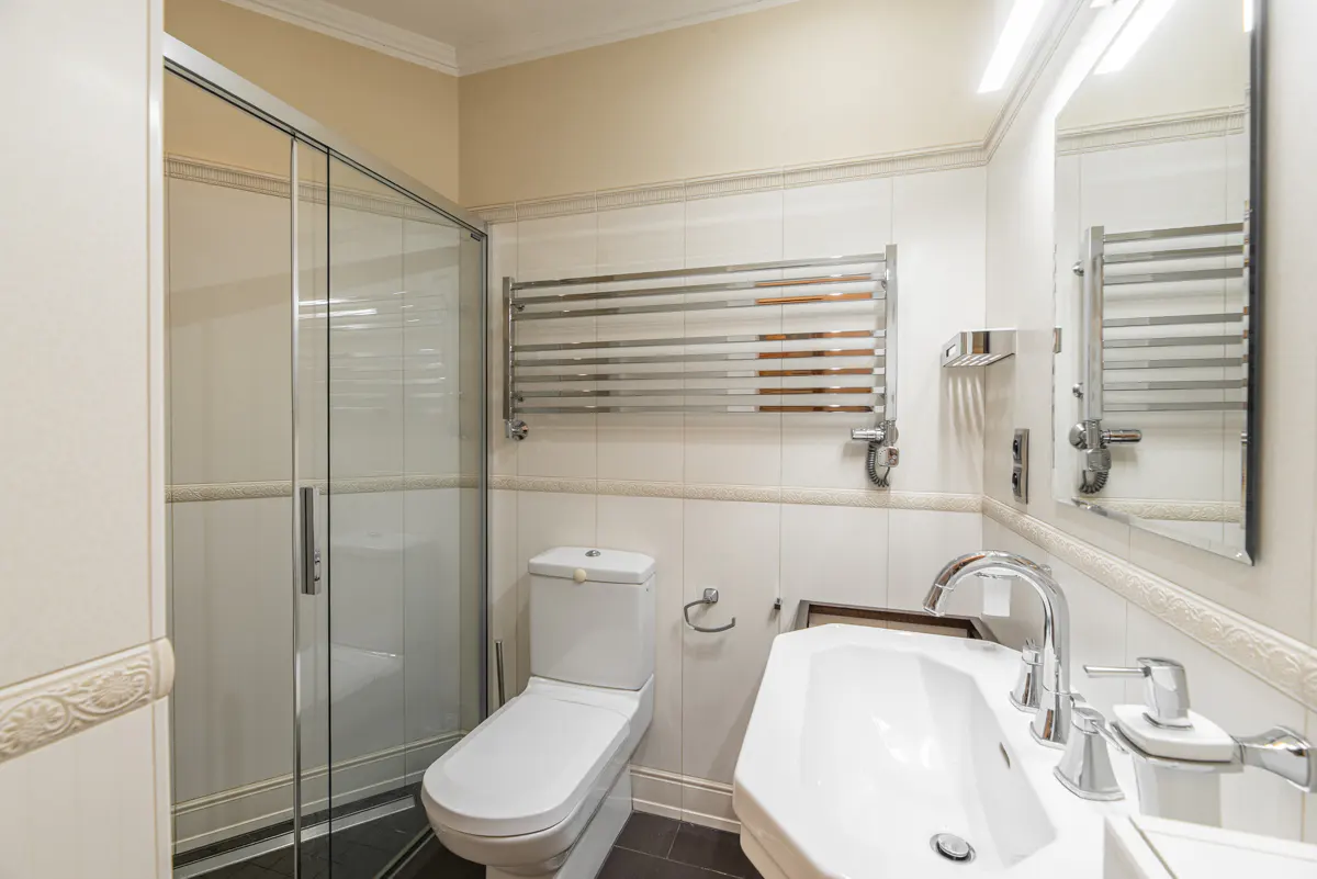 Bright bathroom with white toilet, sink, and tiled walls. A glass shower door and chrome towel rack add modern touches.