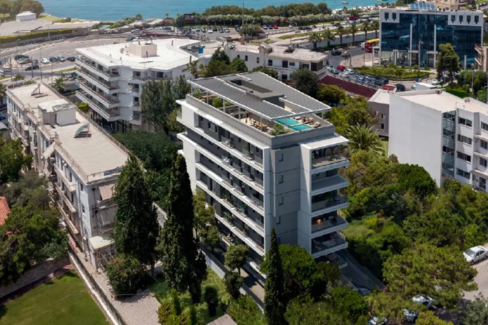 Aerial view of a modern gray apartment building with balconies and a rooftop pool, surrounded by green trees and city buildings.