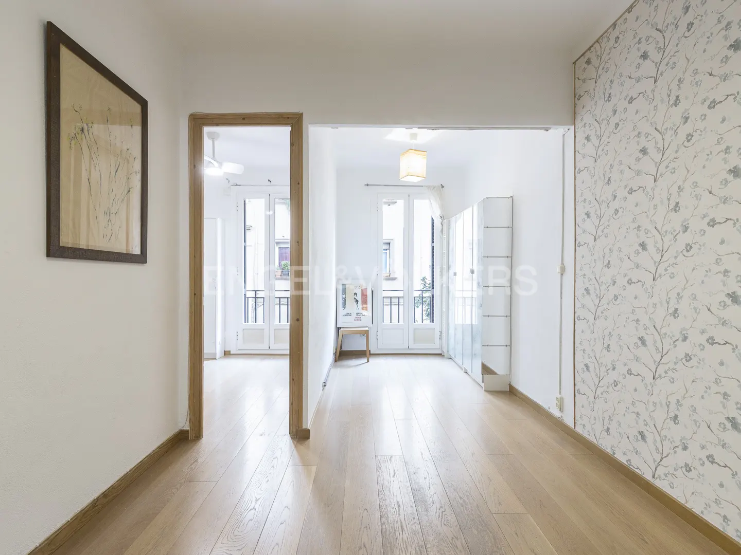 Hallway with light wood floors, white walls, and floral wallpaper. A framed print hangs on the wall. A doorway leads to a bright room with a balcony.