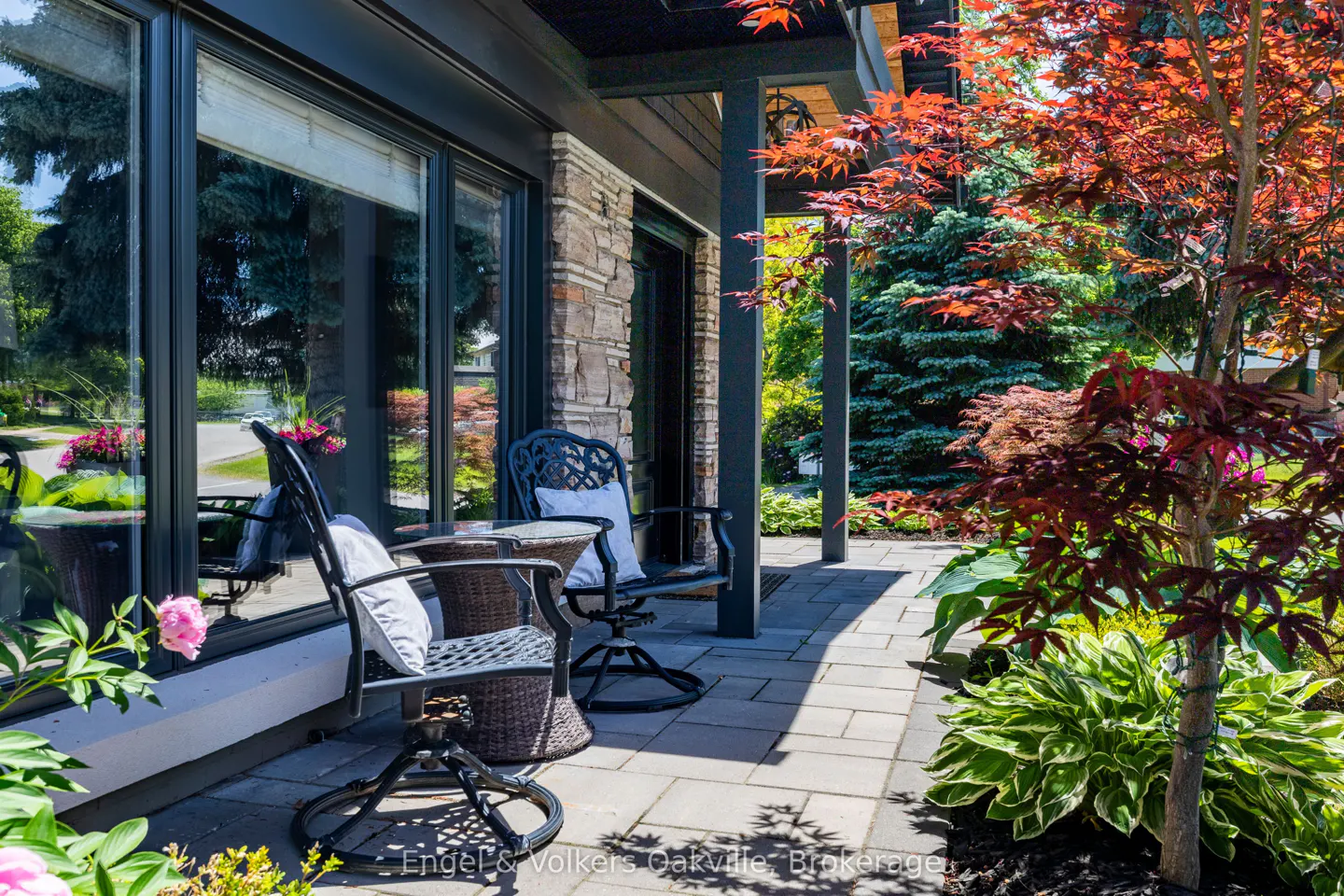 A stone-accented patio features two black metal chairs with white cushions, a small table, and a vibrant red maple tree.