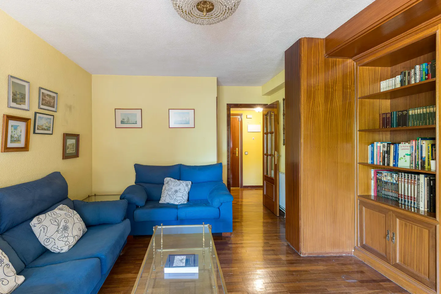 Living room with yellow walls, blue sofas, glass table, and a wooden bookcase filled with books.