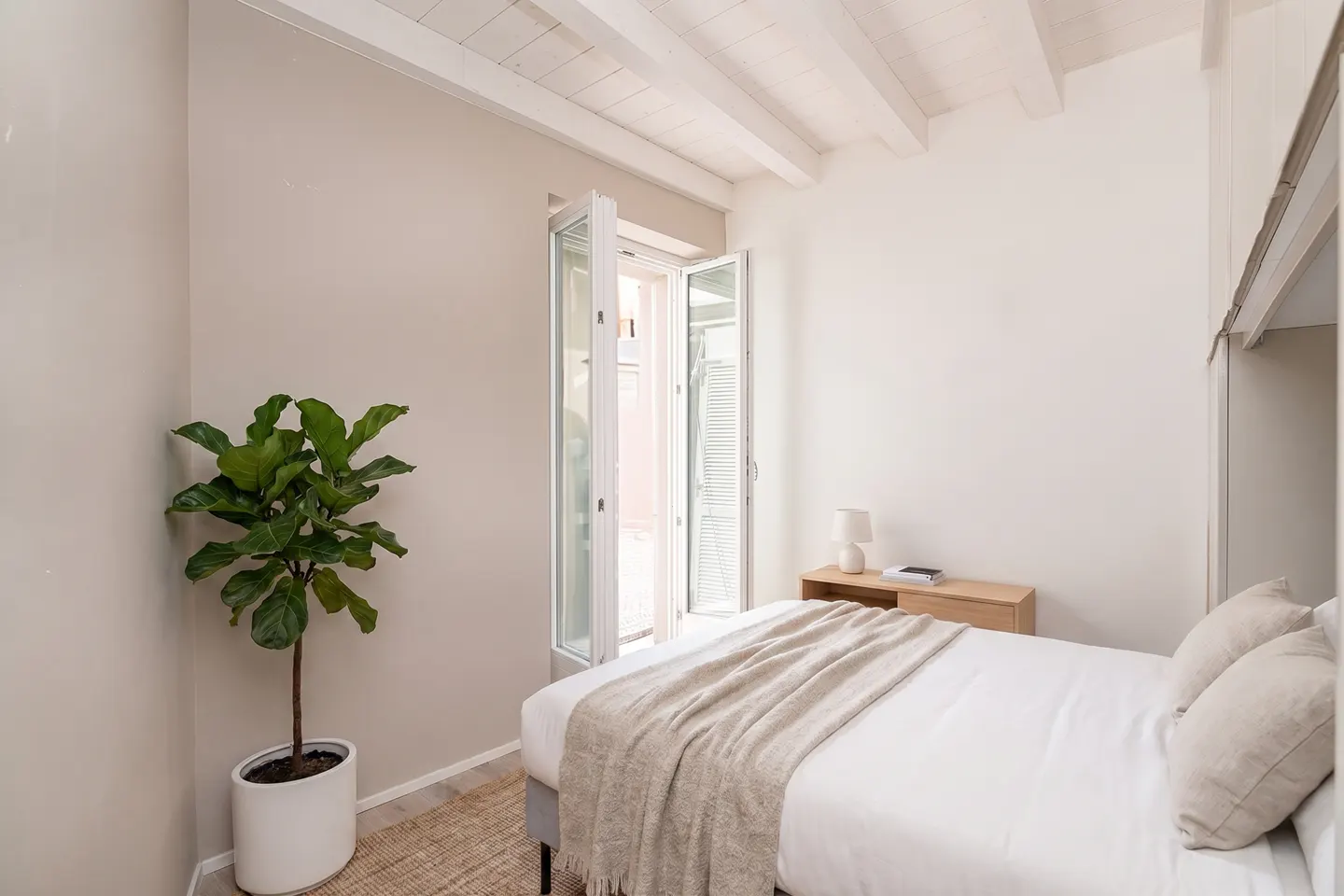 Bright bedroom with white walls, exposed ceiling beams, a double bed, a potted plant, and a balcony door.