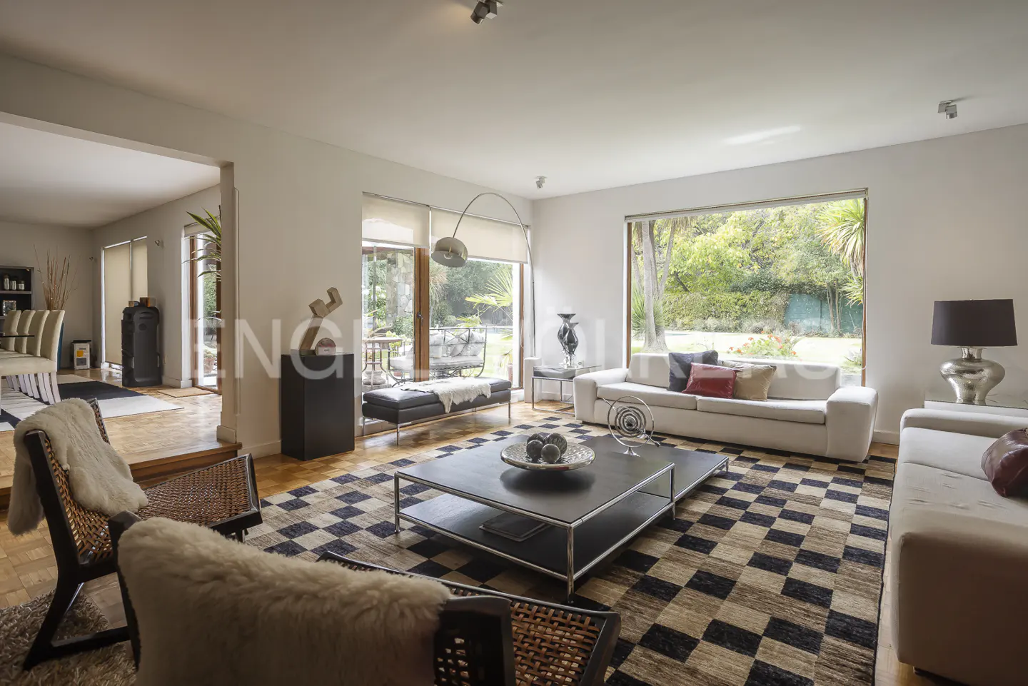 Bright living room with white sofas, a black and white checkered rug, and a large window overlooking a green garden.