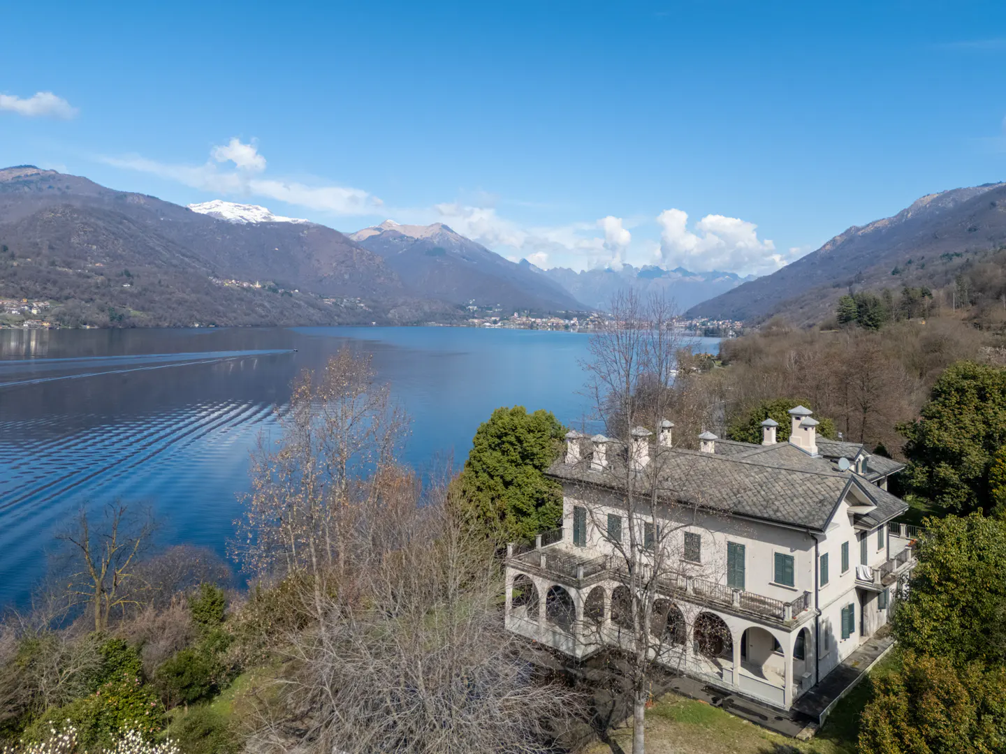 Aerial view of a two-story beige house with a gray roof, overlooking a blue lake and mountains under a clear blue sky.