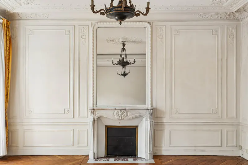 Ornate white room with wood floors, featuring a marble fireplace with a mirror above, reflecting a chandelier. Gold curtains on the left.