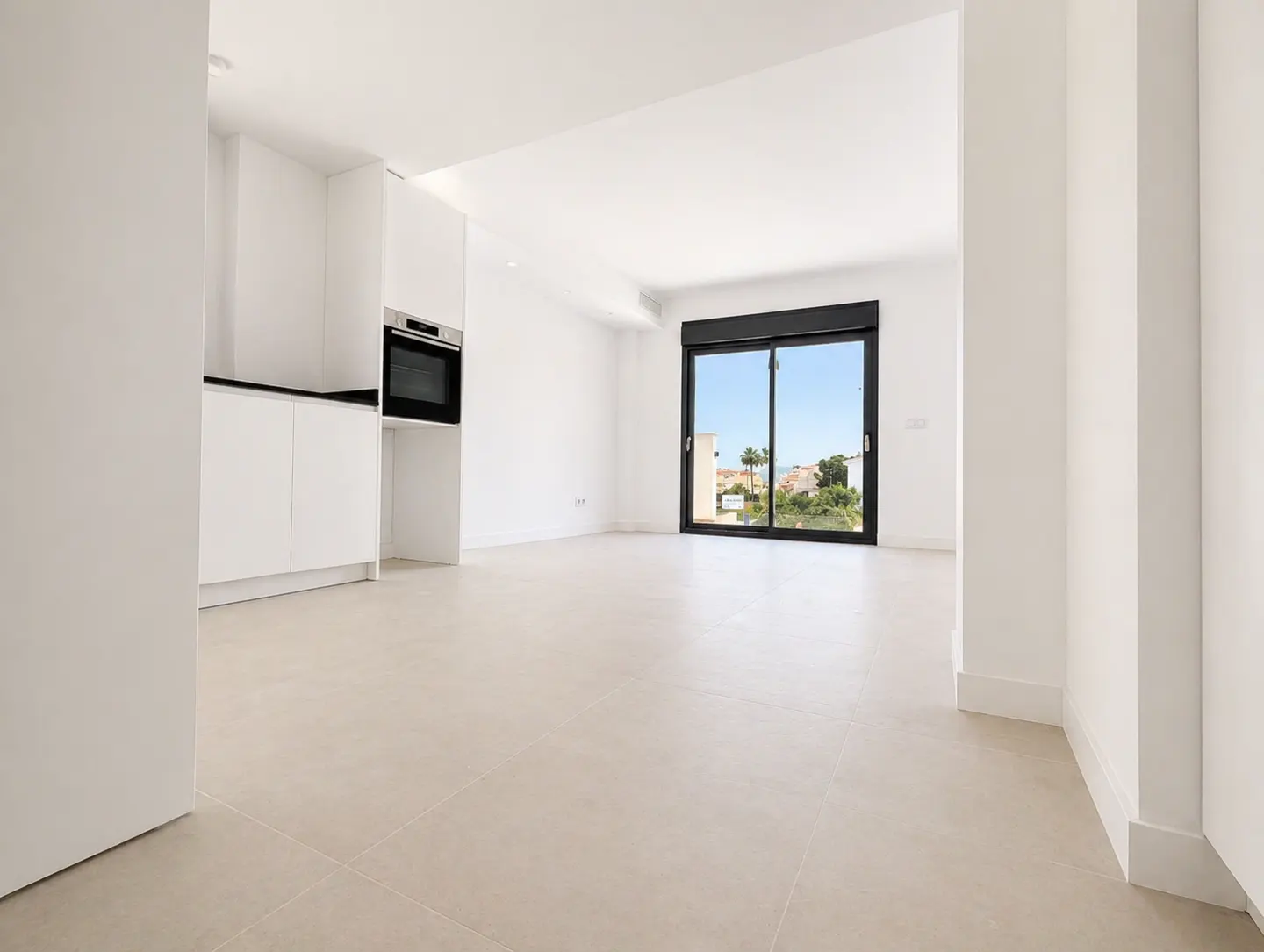 Bright, empty apartment with white walls, tile floor, and kitchen area. A large window shows a view of buildings and trees.