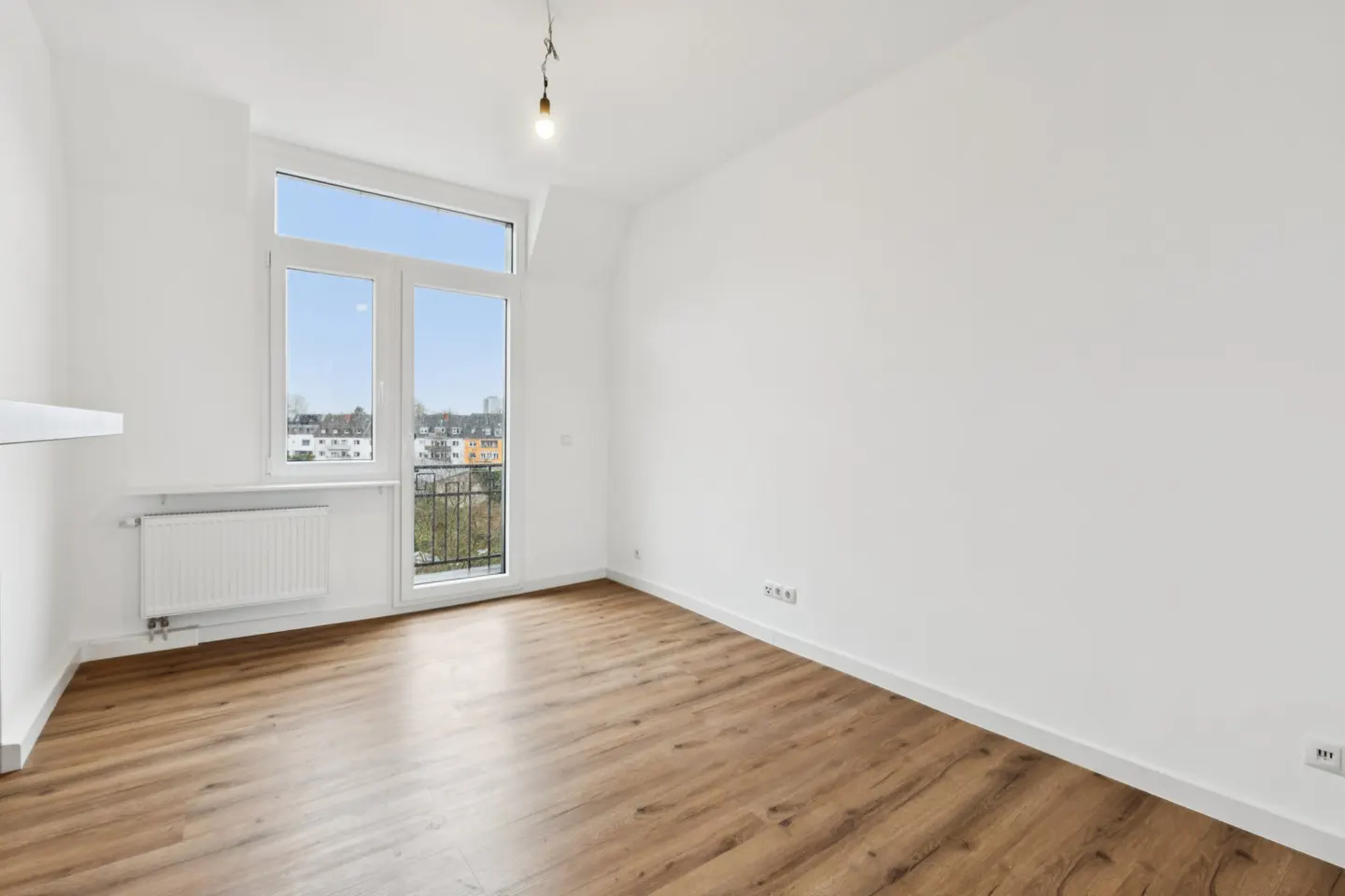 Bright, empty room with wood floors, white walls, and a large window showing a city view. A radiator sits under the window.