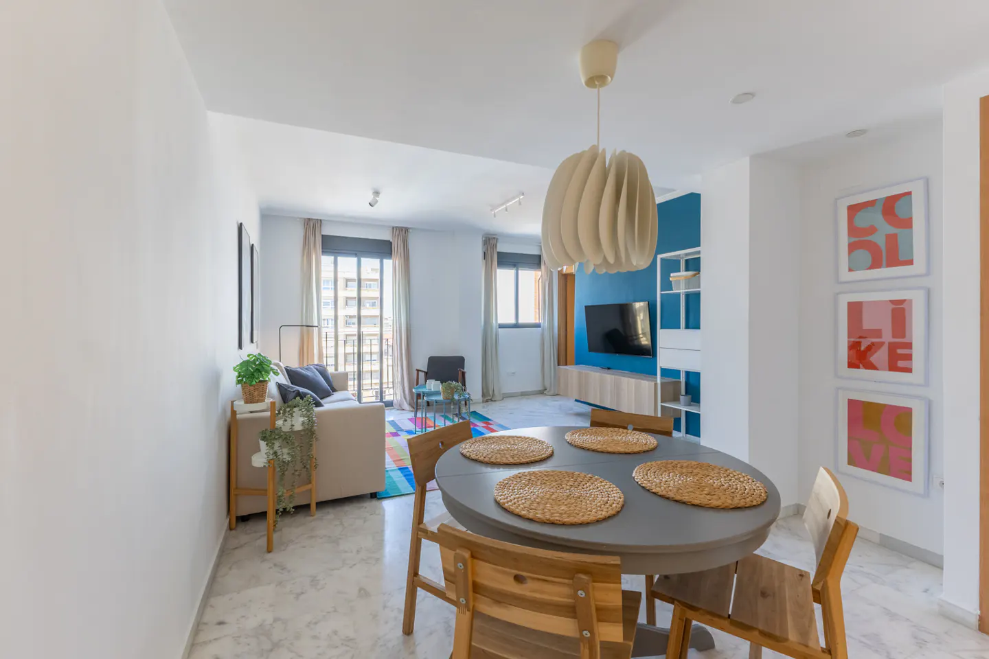 Bright apartment interior with a round dining table, wooden chairs, and a modern light fixture. Living room with sofa and TV in the background.