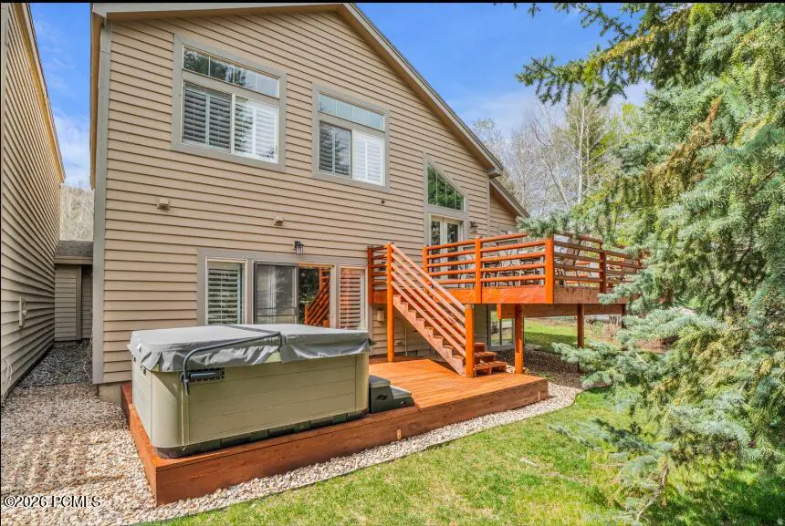 Tan two-story house with a wooden deck, stairs, and a hot tub on a green lawn. Windows with white shutters are visible.