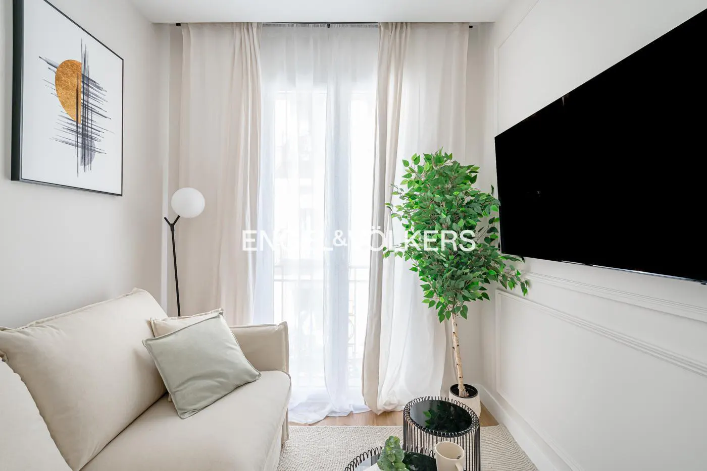 Bright living room with a beige sofa, a black TV, and a green plant near a window with white curtains.