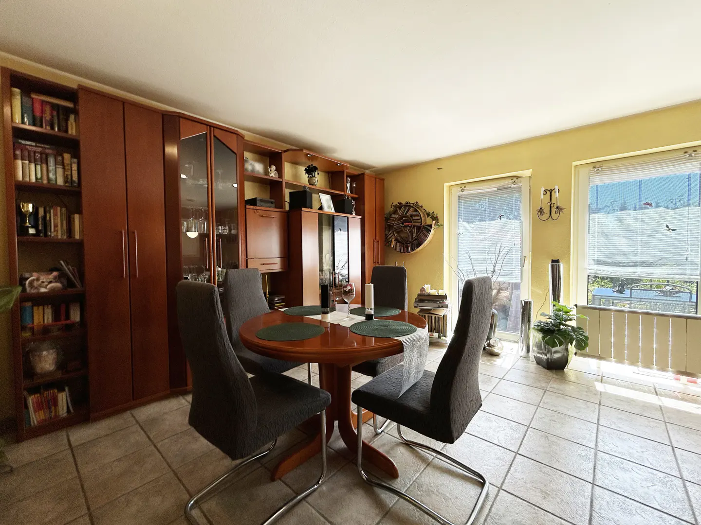 A dining room with a round wooden table, four gray chairs, and built-in wooden cabinets and shelves. Two windows with blinds are visible.