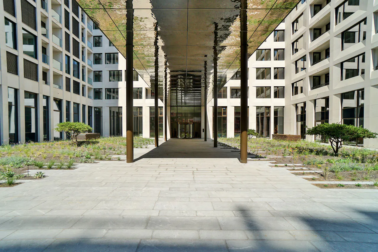 Courtyard view of a modern building with a mirrored canopy, stone walkway, and greenery.