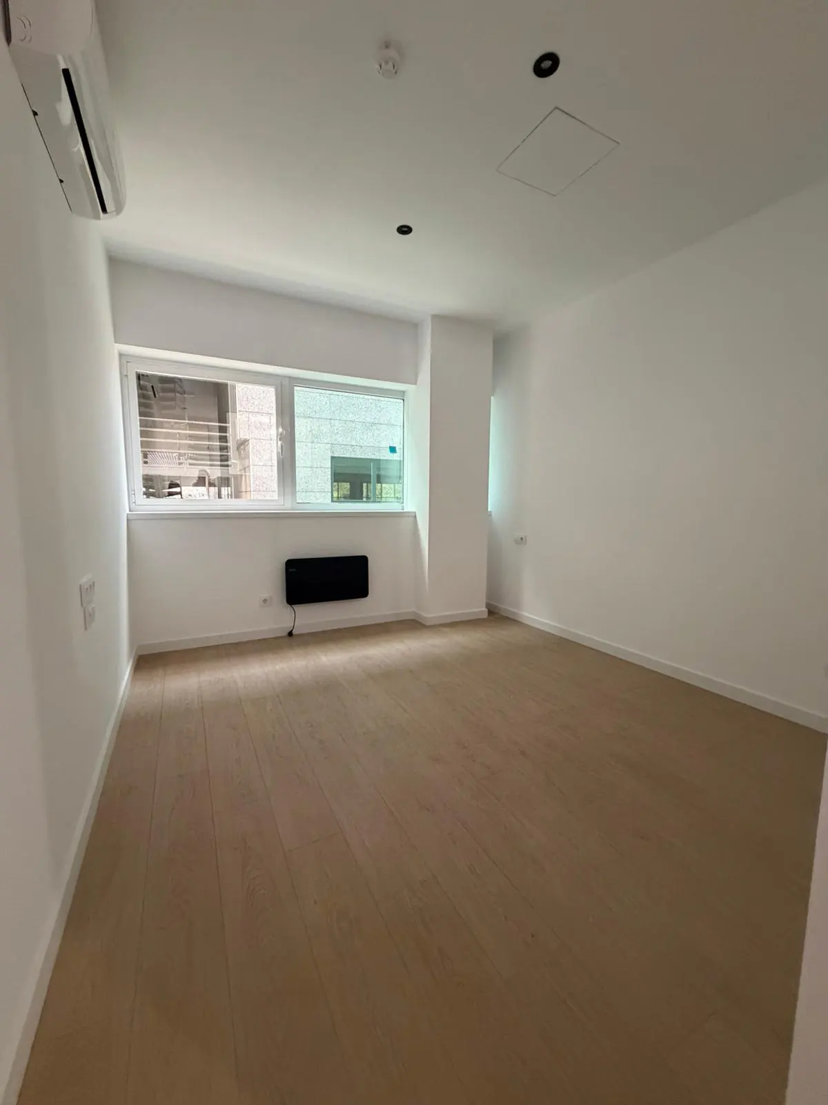 Bright, empty room with light wood floors, white walls, and a window. An air conditioner is mounted high on the wall.