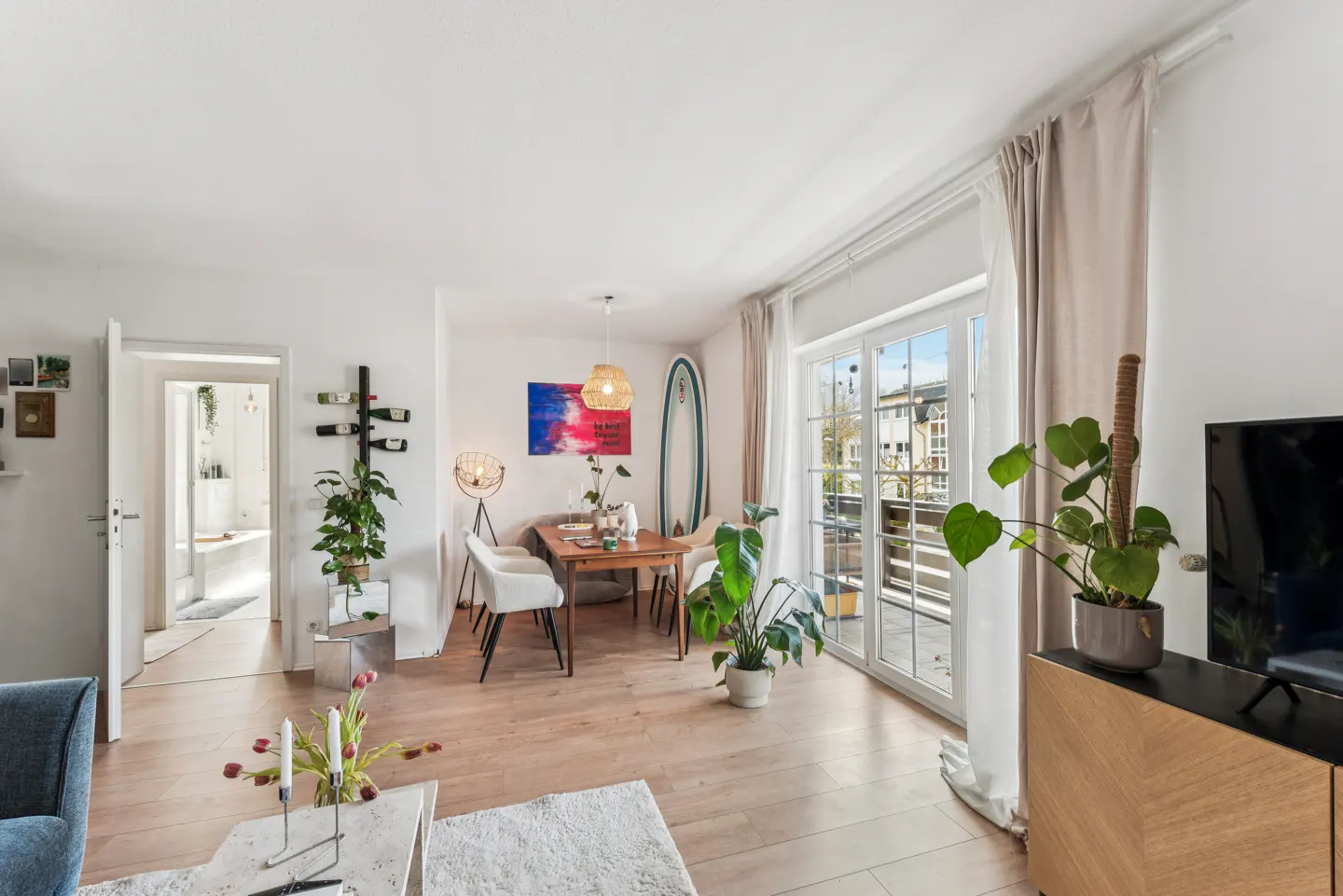 Bright living room with wood floors, white walls, and a dining table. A surfboard and plants add a touch of nature. Balcony doors let in natural light.