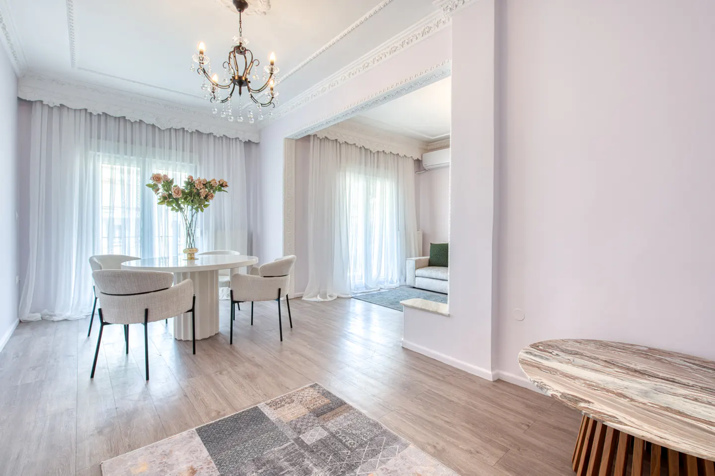 Bright, open-plan living space with white walls, light wood floors, and sheer curtains. A round table with chairs sits under a chandelier.