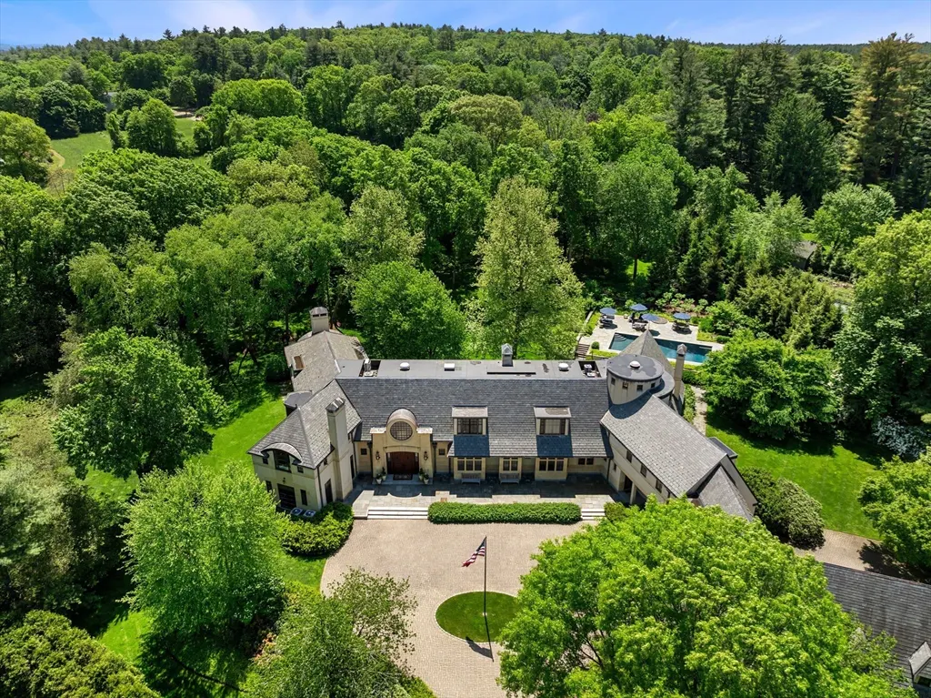 Aerial view of a large, beige house with a gray roof, surrounded by green trees and lawn. A pool is visible in the background. An American flag stands in the front yard.