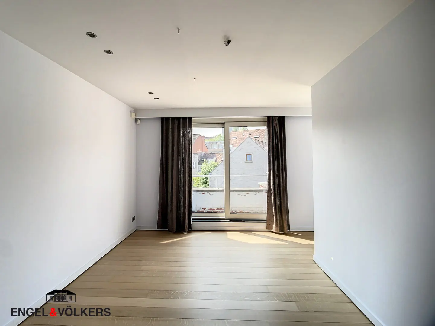 Empty room with light wood floors, white walls, and brown curtains framing a sliding glass door to a balcony.