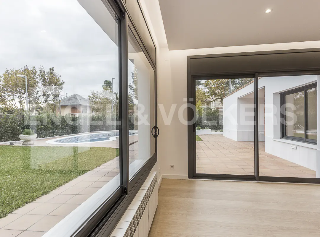 Bright room with wood floors and black framed sliding glass doors. Outside is a pool, green grass, and a white building with a patio.