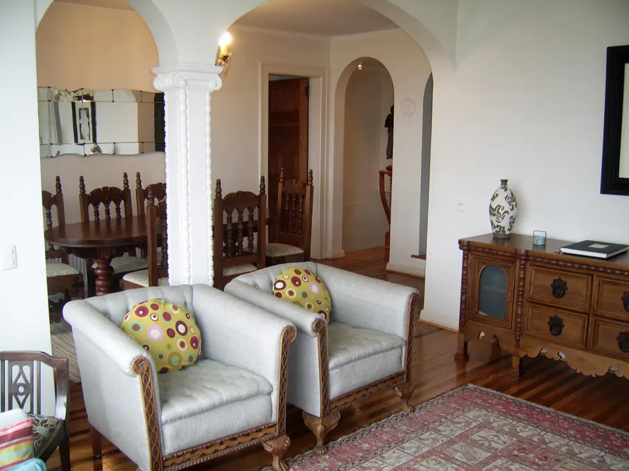 Living room with hardwood floors, two light blue armchairs with colorful pillows, and a wooden dresser with a vase on top. Arched doorways lead to other rooms.