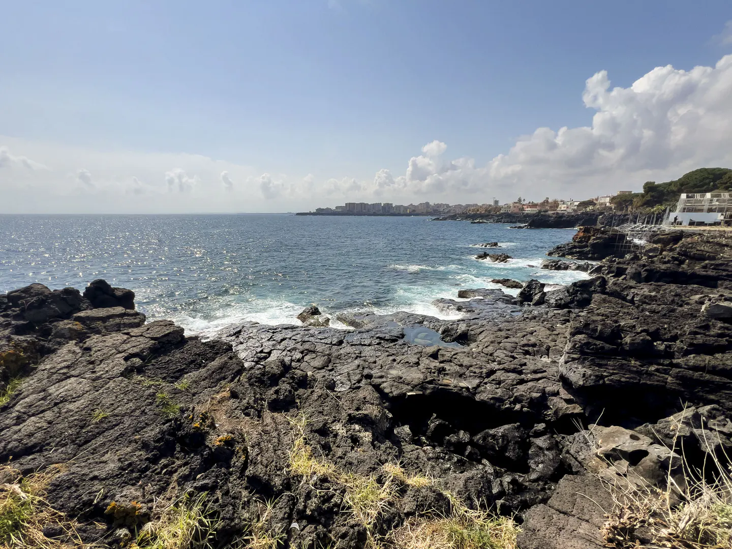 Ocean view from rocky shore. Dark rocks in foreground, blue sea with waves, and a town in the distance under a blue sky with clouds.