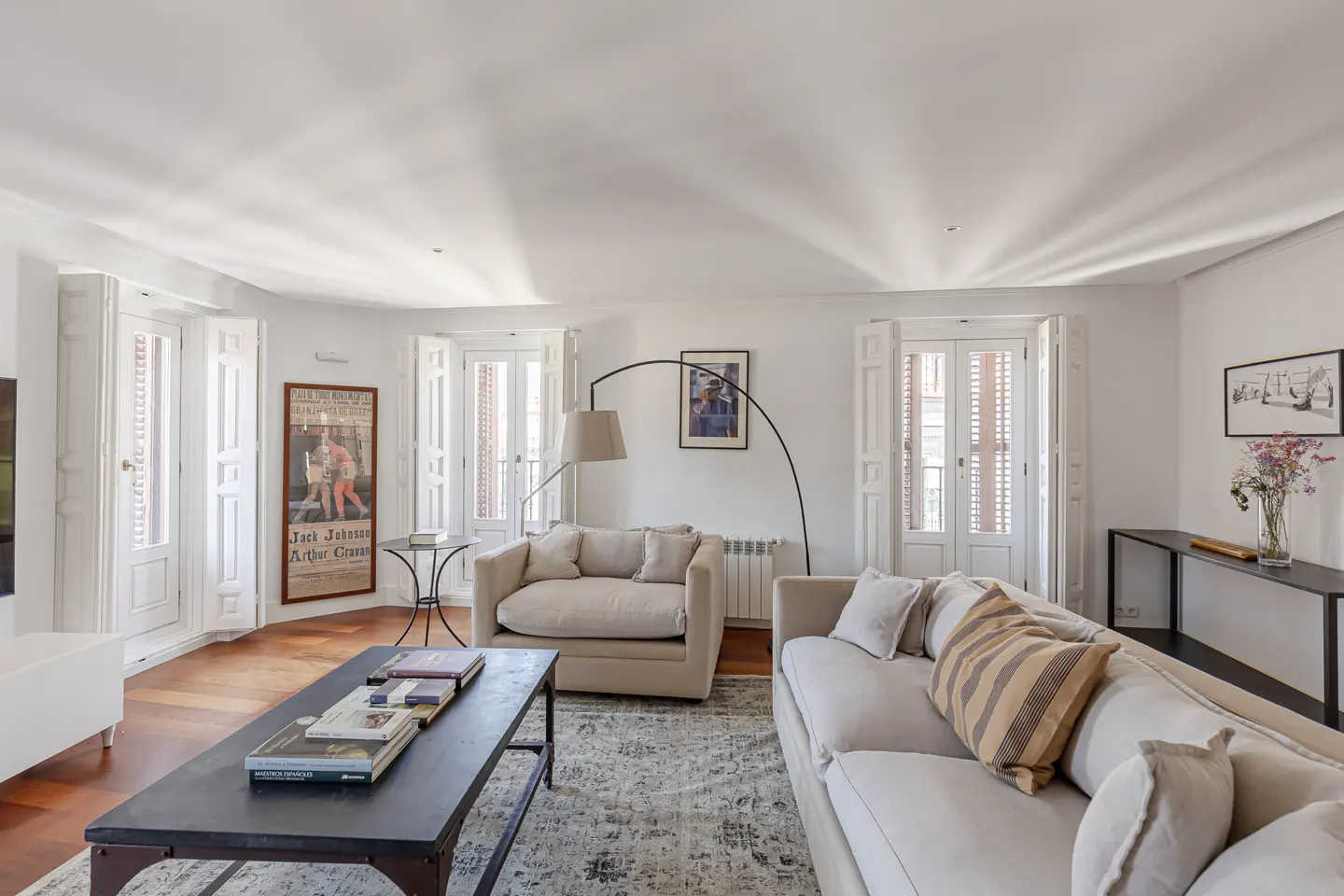 Bright living room with white walls, wood floors, and shuttered windows. Beige sofas, a dark coffee table with books, and a patterned rug create a cozy space.
