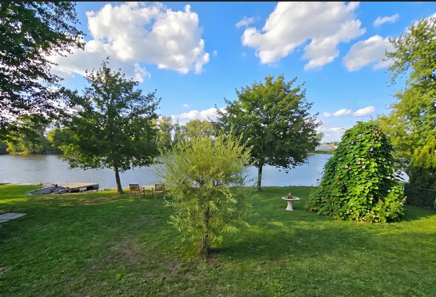 Lush green lawn with trees, a birdbath, and a river view under a blue sky with fluffy white clouds.