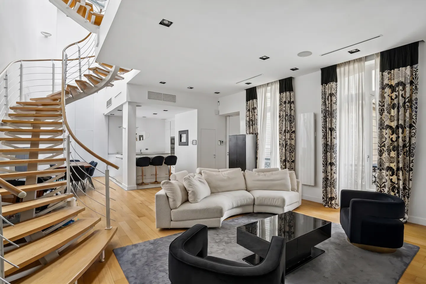 Bright living room with a spiral staircase, white sofa, black chairs, and patterned curtains. Hardwood floors and a glimpse of the kitchen.