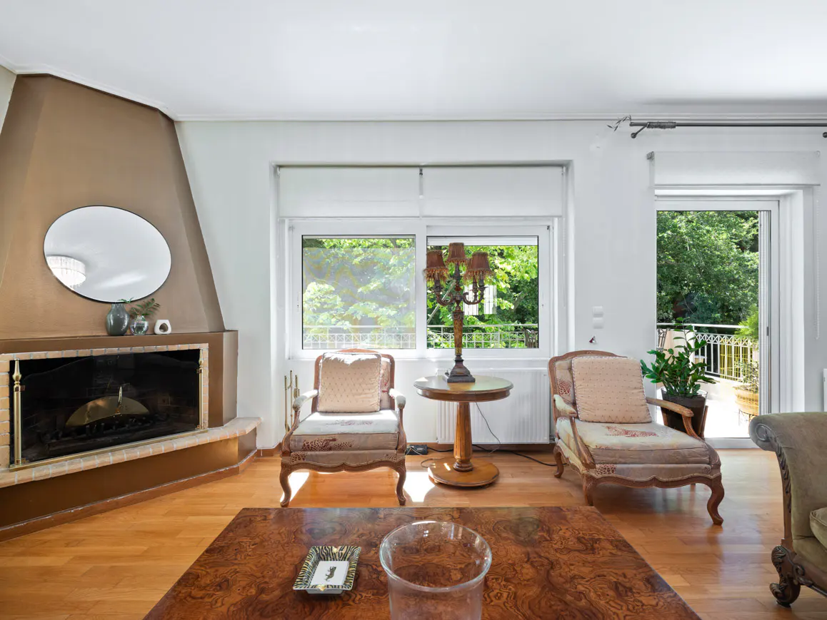 Living room with a fireplace, two armchairs, a wooden side table with a lamp, and a view of a balcony with greenery.