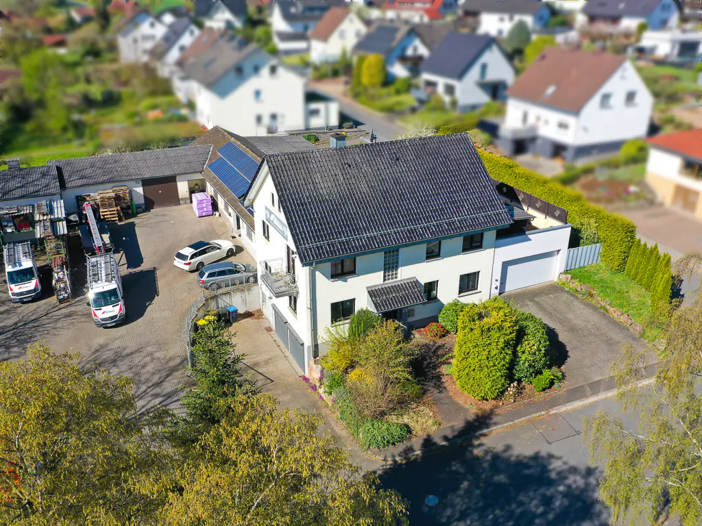 Aerial view of a two-story white house with a dark gray roof, surrounded by green trees and shrubs. Several vans and cars are parked nearby.