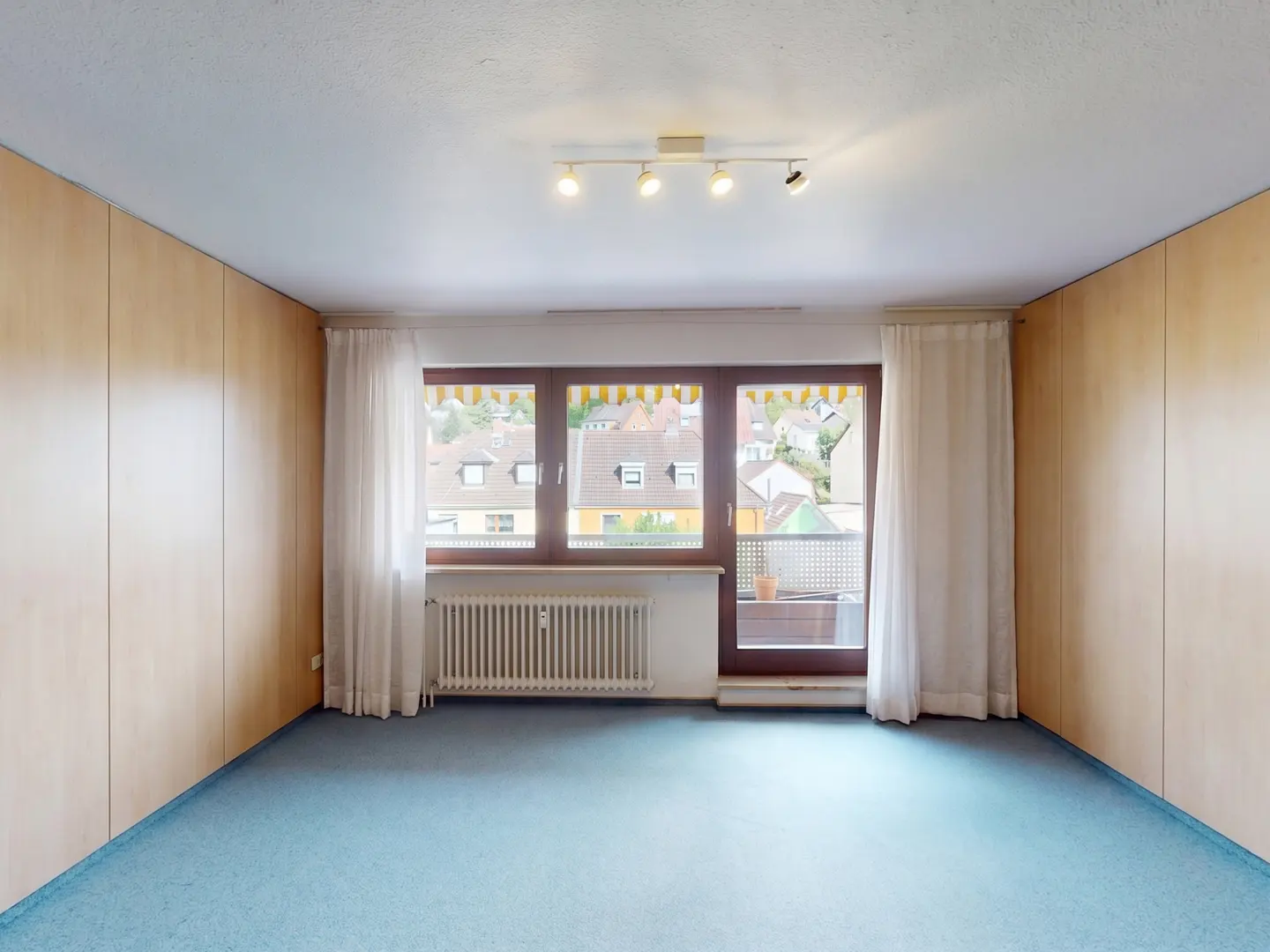 Empty room with light wood paneled walls, blue carpet, white curtains, a radiator, and a balcony door overlooking houses.