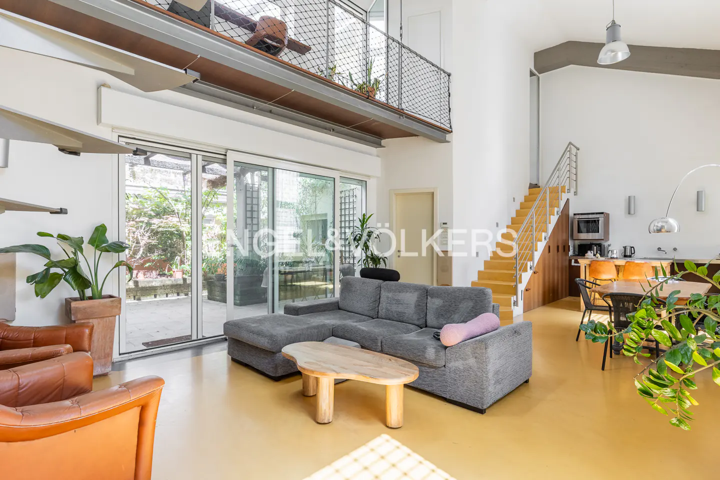 A modern living room with a gray sofa, wooden table, and orange chairs. A staircase leads to a loft with a metal railing. Large windows open to a patio.
