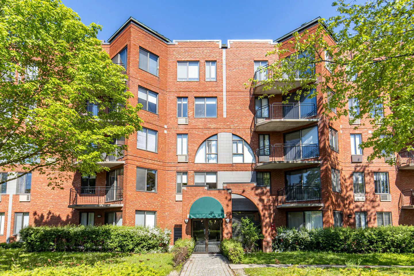Brick apartment building with balconies, green awning over the entrance, and trees on either side.