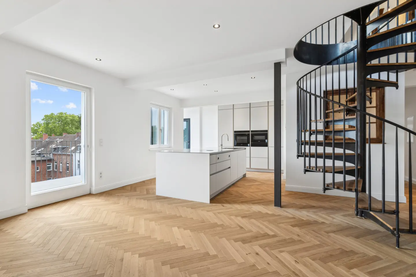 Bright, modern apartment interior with herringbone wood floors, white walls, kitchen island, and a black spiral staircase. A large window shows a cityscape.