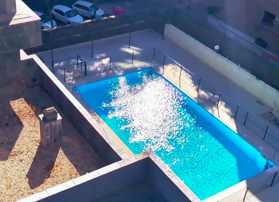 Aerial view of a rectangular, bright blue swimming pool surrounded by a fence and concrete patio. Sunlight glints off the water.