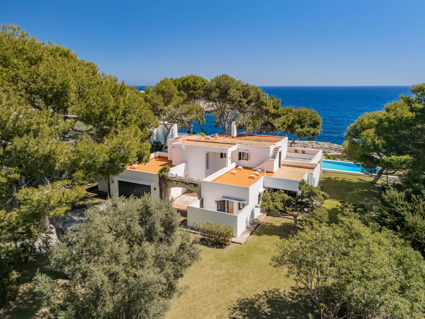 Aerial view of a modern white house with orange roof, surrounded by green trees, with a blue pool and ocean in the background.