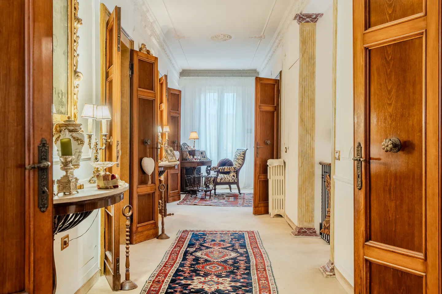 Hallway with wood doors, columns, and a patterned rug. A desk and chair are visible in the background.