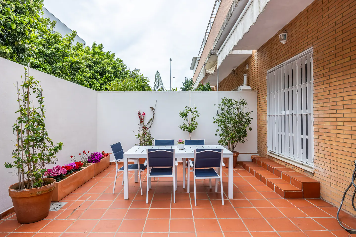 Outdoor patio with white table and blue chairs on terracotta tile. White walls, greenery, and a brick building are visible.