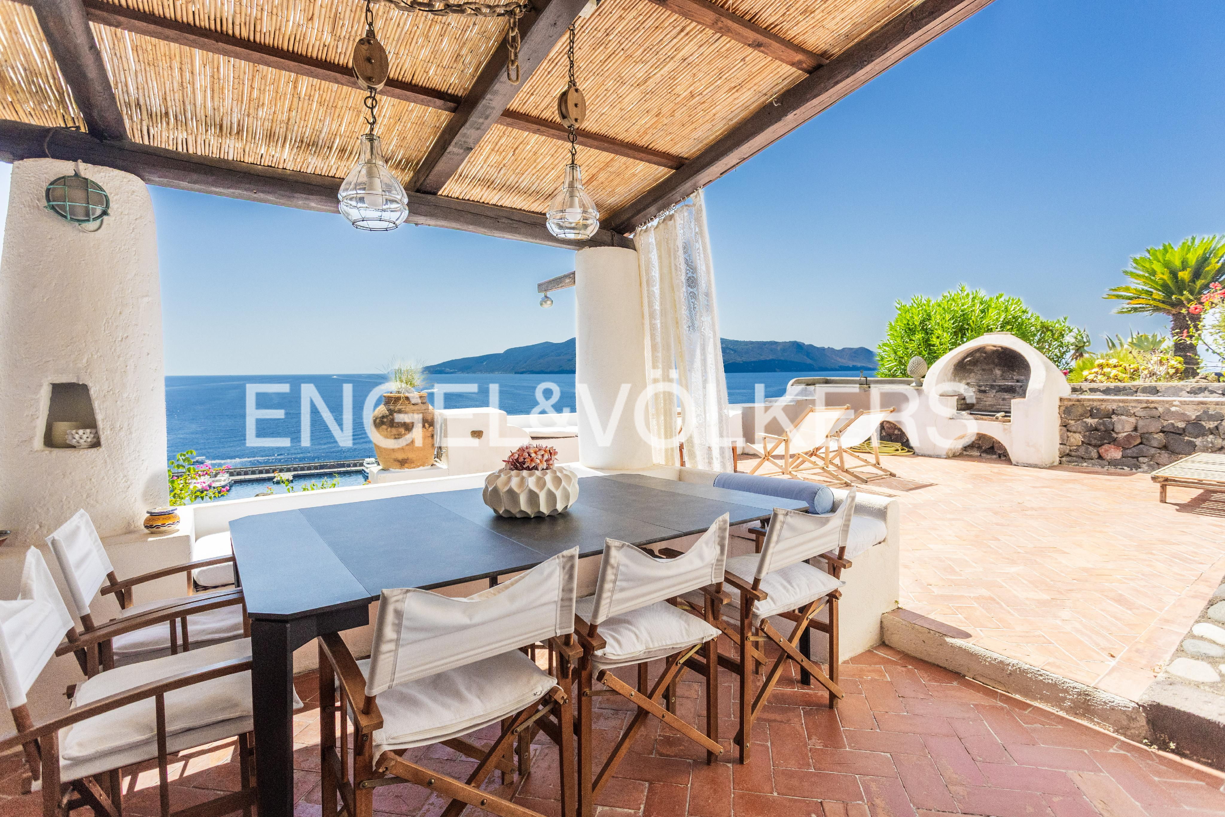 Outdoor dining area with a dark table and white chairs under a thatched roof, overlooking the blue sea and green landscape.