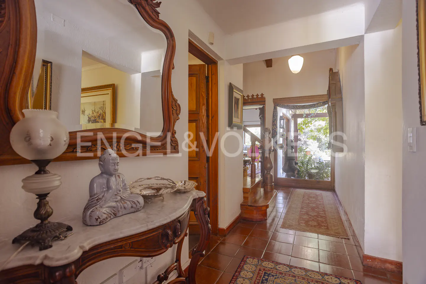 Foyer view with a wooden console table, lamp, and Buddha statue. A large mirror reflects a framed painting. The hallway leads to a garden view.