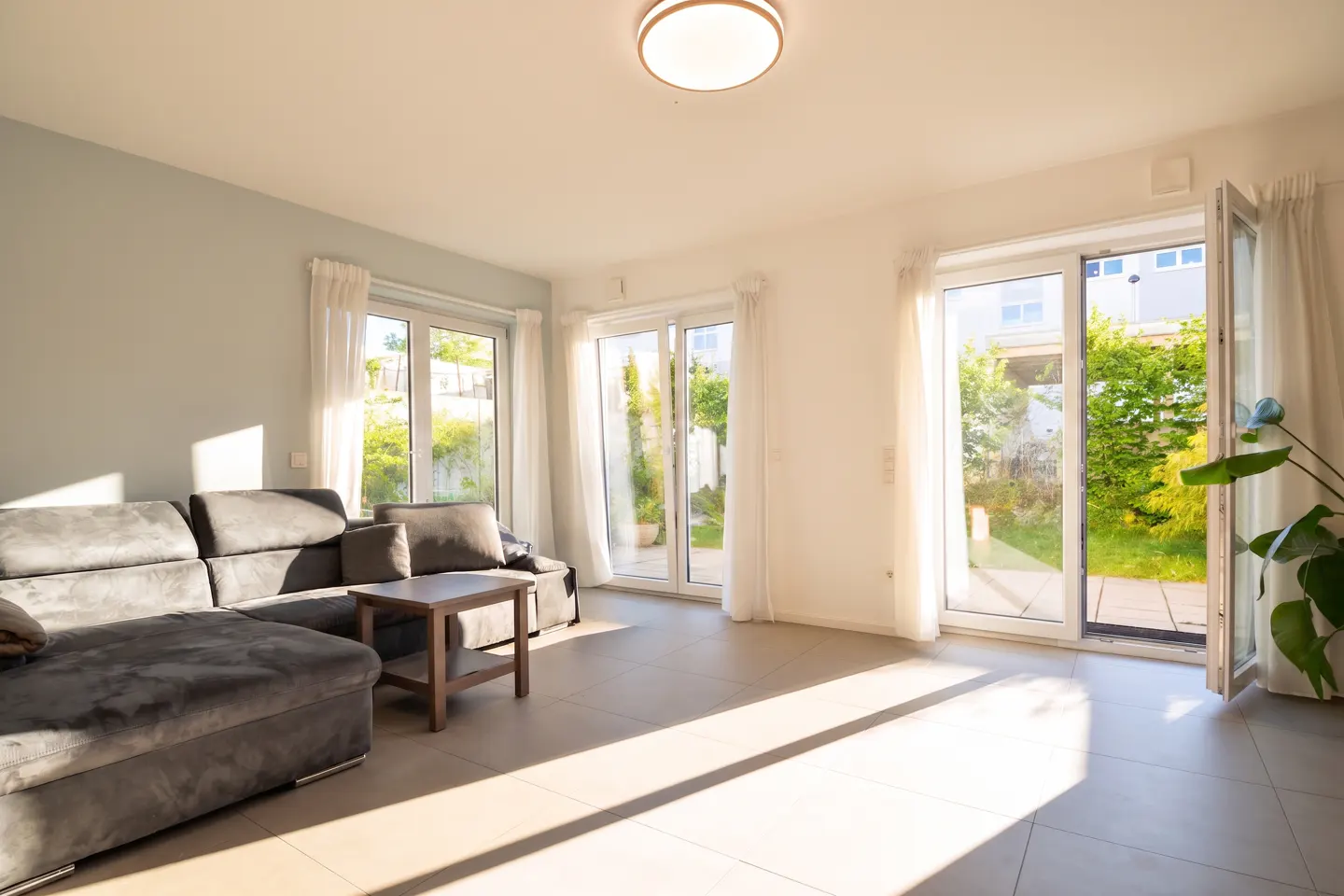 Bright living room with a gray sectional sofa, wood table, and sliding glass doors to a green yard. Sunlight streams in.