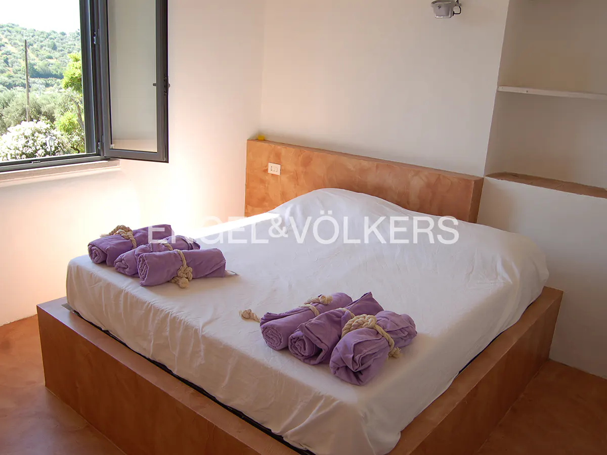 Bedroom with a white bed, a wooden headboard, and purple towels. An open window shows a green landscape.