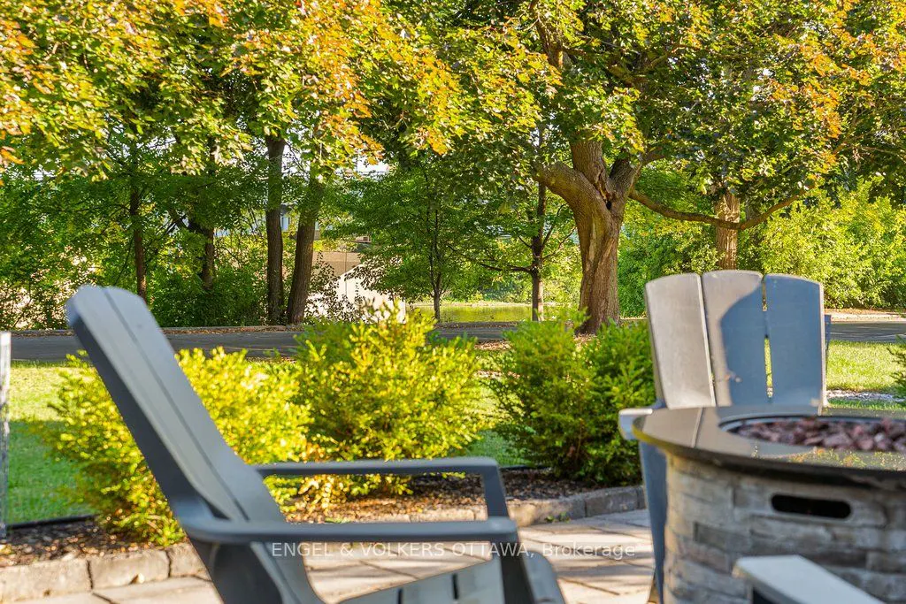 Outdoor patio with gray chairs and a stone fire pit, surrounded by green bushes and trees.
