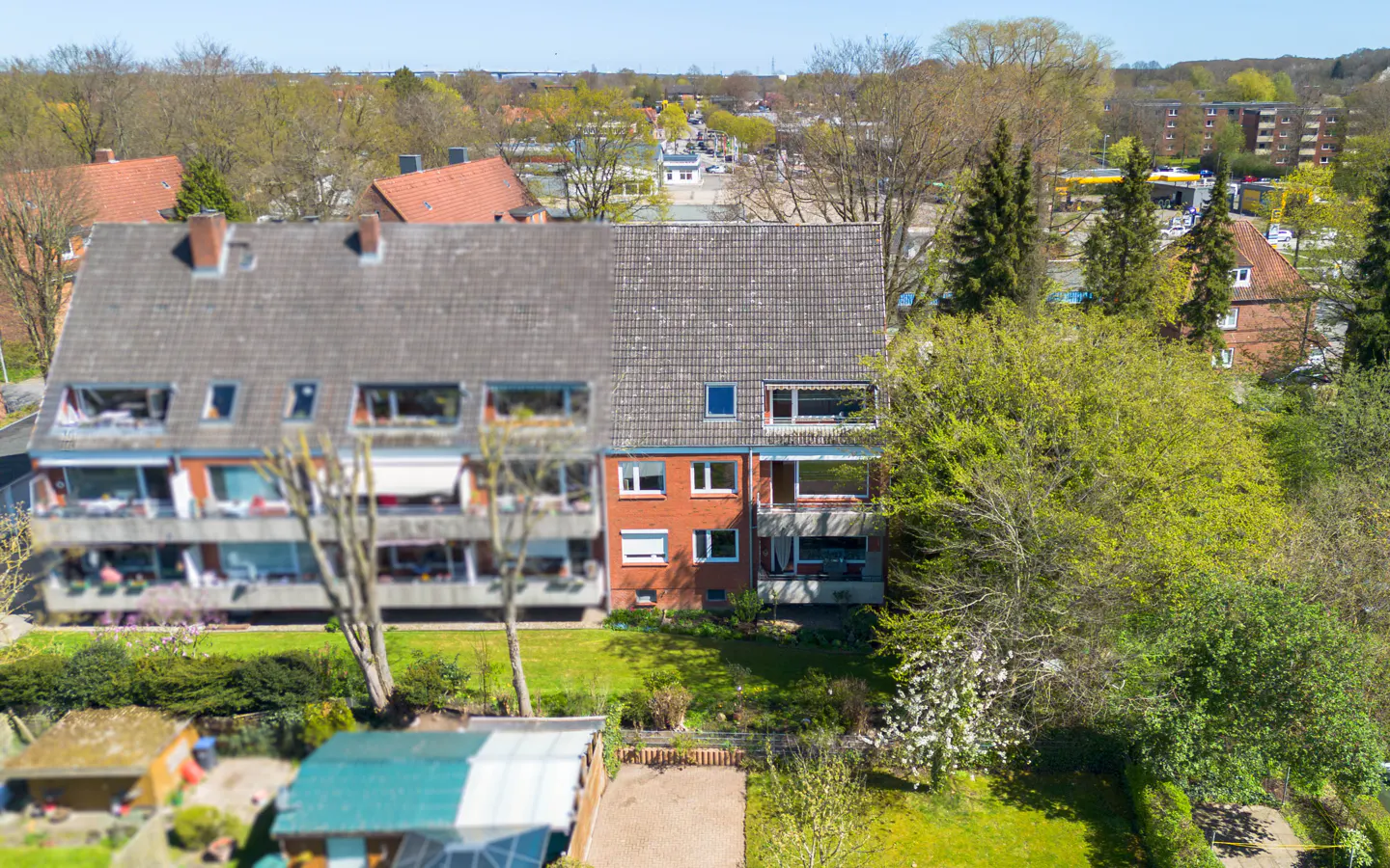 Aerial view of a multi-story apartment building with balconies, a gray roof, and a red brick facade, surrounded by green trees and lawns.