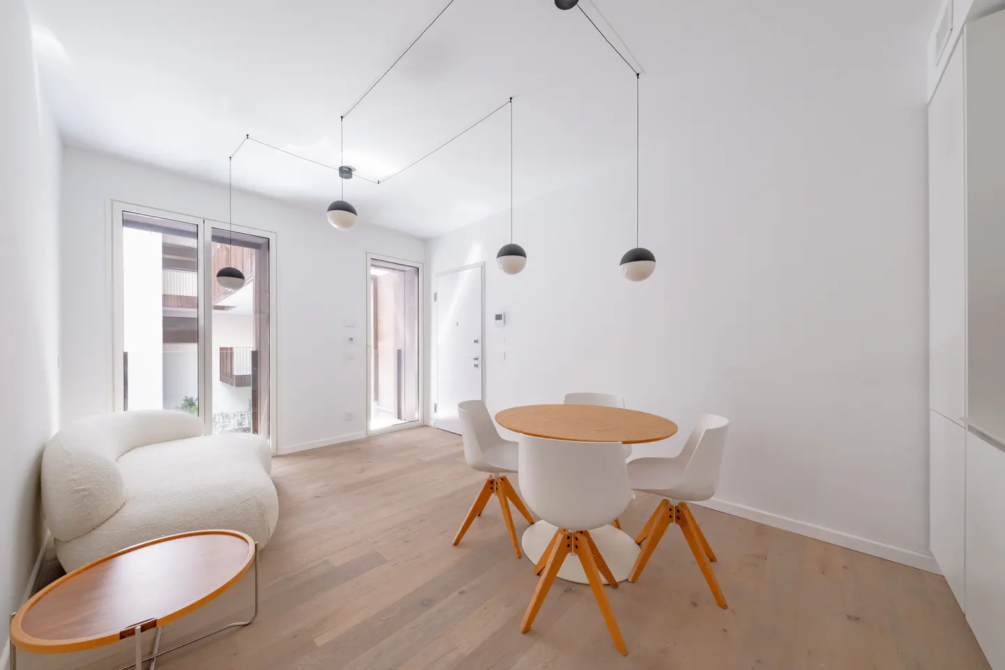 Bright, modern apartment interior with white walls, wood floors, and minimalist furniture. A round wood table with white chairs sits under globe pendant lights. A white sofa is near a wood-framed glass door.