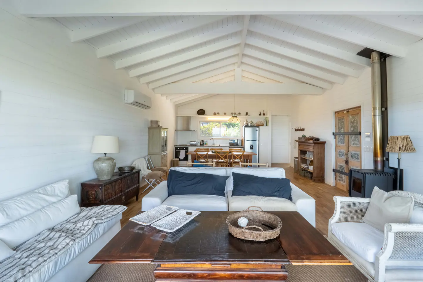 Bright living room with white walls, vaulted ceiling, wood stove, and open kitchen in the background.
