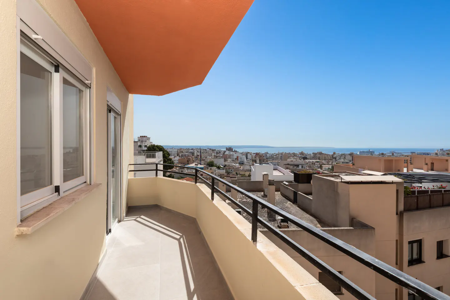 Balcony view of city. Beige walls, black railing, and orange ceiling. Cityscape and blue sky in the background.