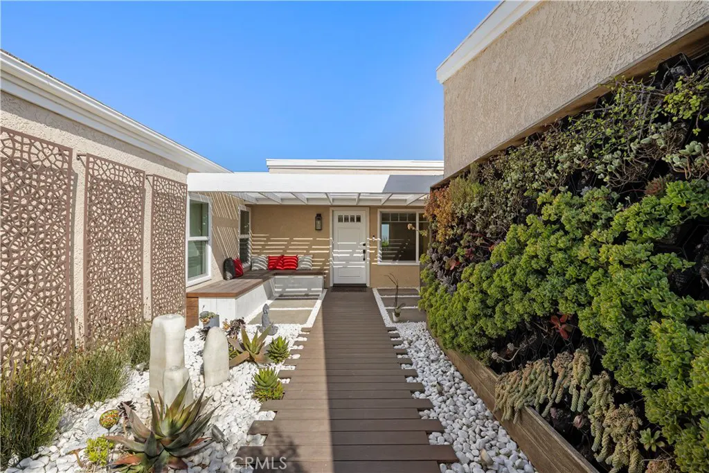Front entrance of a tan house with a white door, a wooden walkway, and a green wall of plants.